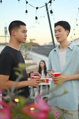 Two young handsome men having drinks and chatting at the rooftop party in the evening.