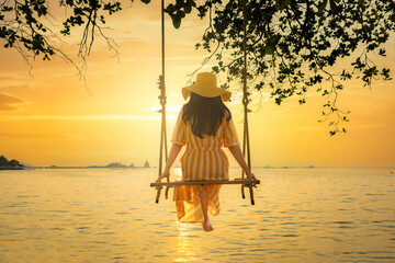 Happy young woman in yellow dress and hat sitting on swing enjoy freedom and summer on sea shore on sunset. Beautiful golden sunlight reflection with ripple on sea surface in sunset time.