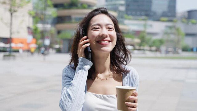 Charming young asian businesswoman talking on the mobile phone happy chatting online in the sunny urban squre with coffee cup in hand