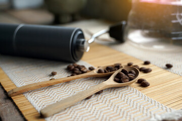 Roasted coffee beans in a wooden spoon and a coffee grinder on a woven bamboo placemat. It is a...