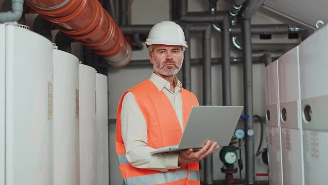 Caucasian factory worker in white hard hat and orange safety vest typing on modern digital laptop at workplace. Gray haired man looking at camera with serious face while managing technological process
