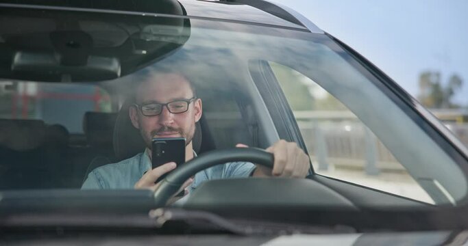 Young Caucasian man sits in a car on a parking lot and typing message on a smart phone