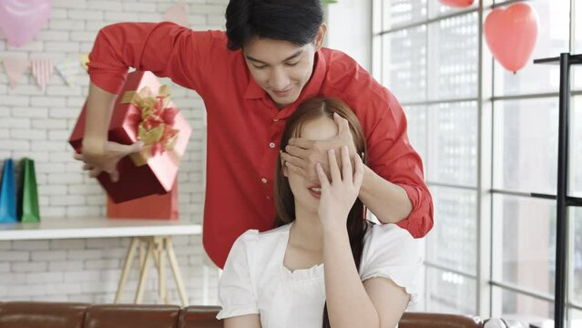 Smiling young asian husband or boyfriend closes eyes to his wife or girlfriend and gives gift box in living room interior. Birthday surprise, valentine day and anniversary, romantic date at home 