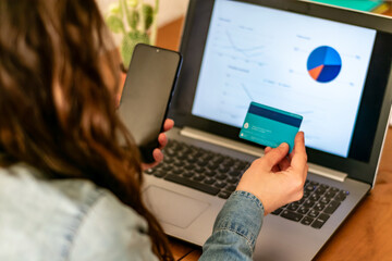 professional woman is sitting at her desk at home, working on her laptop. She is holding a bank card in her hand and checking statistics on her computer. She is making an Internet transaction.