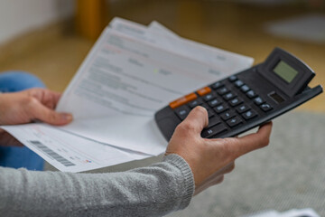 A young entrepreneur is sitting on the floor of her home, calculating bills. She has a calculator in her hand.