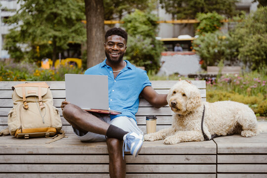 Portrait of smiling man with laptop sitting by labradoodle dog on bench in park