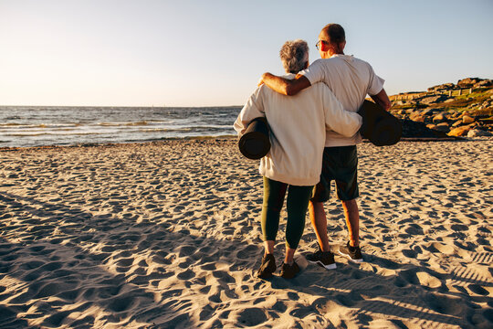 Rear view of senior couple holding exercise mats while standing on sand at beach