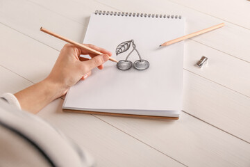 Woman drawing cherries with graphite pencil in sketchbook at white wooden table, closeup