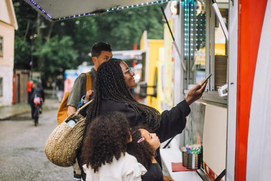 Smiling woman doing online payment near food truck while standing with family at amusement park