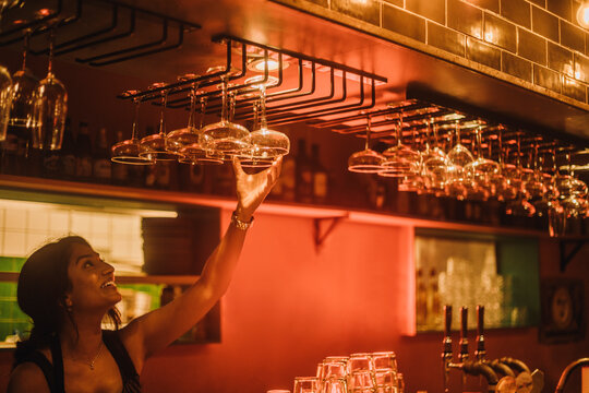 Smiling female bartender hanging cocktail glasses on rack while working at bar