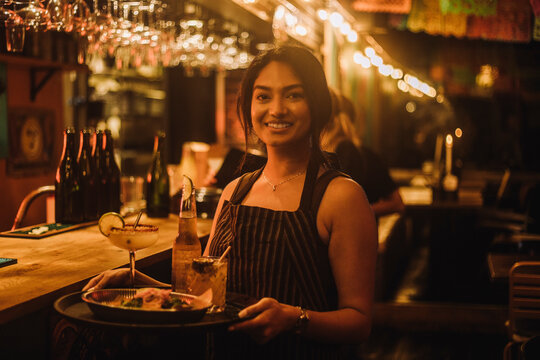 Portrait of smiling waitress holding food and drinks on serving tray while working at bar
