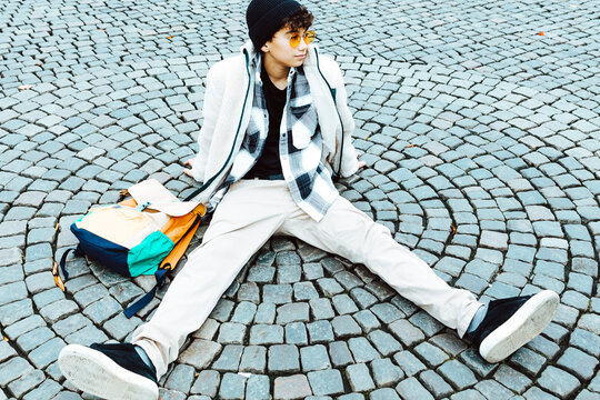 Boy with backpack sitting on footpath while hanging out in city