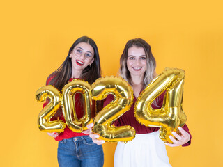 Beautiful women celebrating 2024, happy gorgeous female in casual dress holding gold color 2024 balloons, good times at New Year's Eve party. Holiday celebration. Isolated yellow. Copy space.