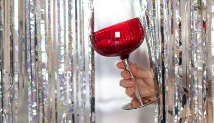 Woman's hand holding red glasses on the silver tinsel foil fringe background. Christmas celebration...