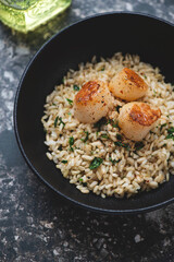 Closeup of seared scallops with brown rice, lemon zest and parsley served in a black bowl, vertical shot, selective focus