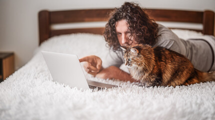 A bearded man with a cat is working on a laptop - obrazy, fototapety, plakaty