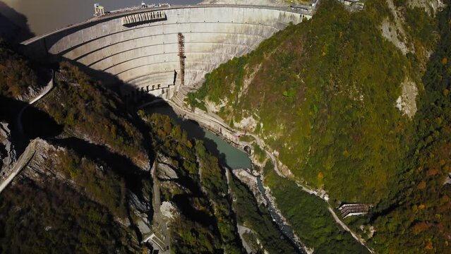 Huge concrete wall of view dam located amidst majestic mountains covered by green forest filmed by drone at summer day. Impressive engineering work in form of the weir. Union of nature and industry