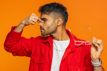 Exhausted tired Indian young man takes off glasses, feels eyes pain, being overwork burnout from long hours working. Sleepy exhausted Arabian guy rubbing eyes isolated on orange background indoors