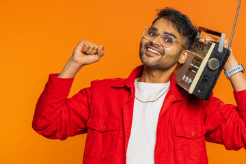 Happy young Indian man using retro tape record player to listen music, disco dancing favorite track, having fun entertaining, fan of vintage technologies. Hindu guy isolated on orange background