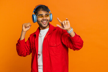 Happy stylish Indian man in sunglasses listening music via headphones, dancing disco fooling around having fun expressive gesticulating hands. Arabian excited guy isolated on orange background indoors - obrazy, fototapety, plakaty