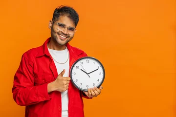 Foto op Canvas It is your time, hurry up. Indian young man showing time on wall office clock, ok, thumb up approve pointing finger at camera, advertisement, deadline. Arabian guy on orange background. Copy-space © Andrii Iemelianenko