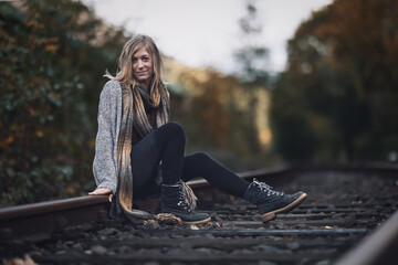 Young woman with long blonde hair and a long knitted coat sits on a railway track and smiles at the...