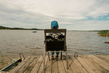 An unrecognizable school-age boy sits alone on a chair on the calm shore of the lake