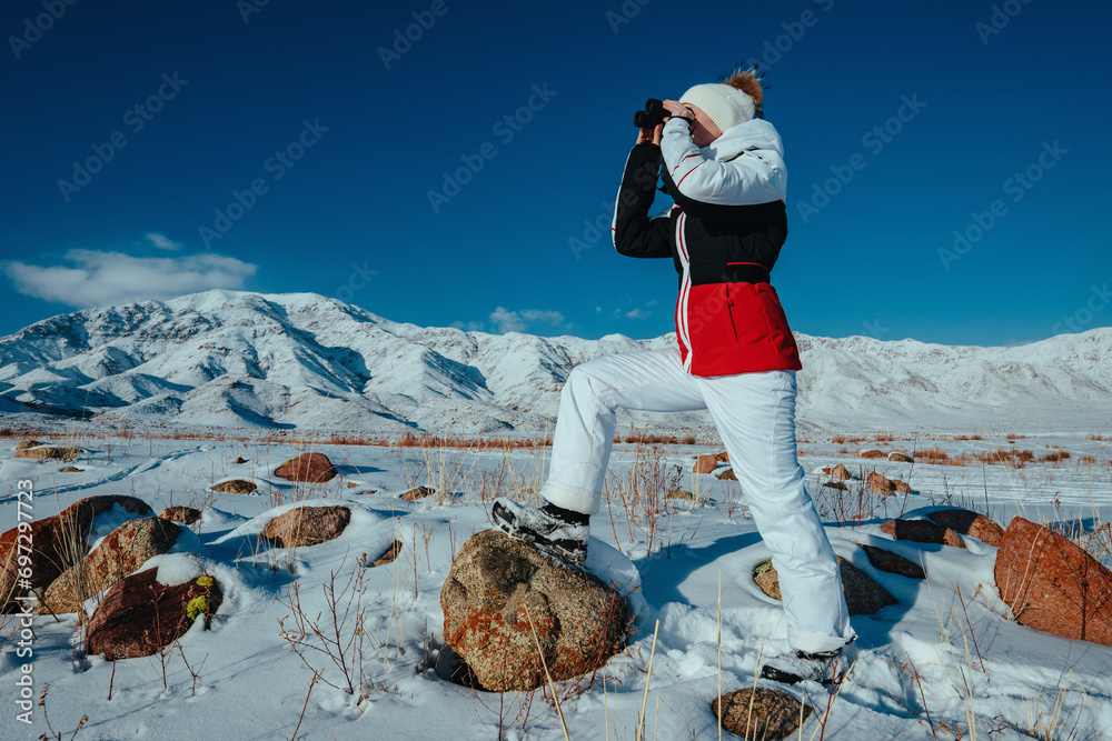 Wall mural Young woman tourist looking through binoculars on mountains background in winter day