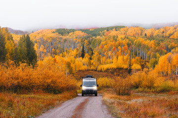 Adventure Travel Vanlife Van Drives Off Road Dirt Dusty Path Through Colorado Forest in Autumn. Colorful Yellow Aspen Trees and Moody Low Clouds Fog