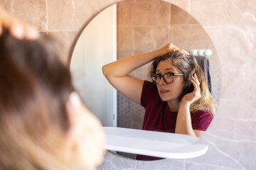 Gray haired surprised caucasian middle aged woman looking at grey hair head in mirror reflection. Early gray hair concept