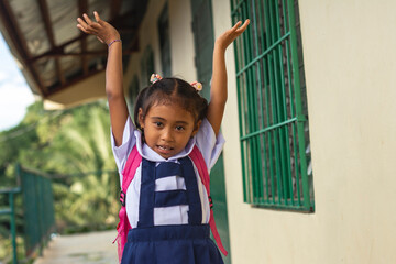 A simple cute preschooler asian girl throws her hands into the air, celebrating while walking outside her classroom. At a provincial primary school in Bohol. - obrazy, fototapety, plakaty
