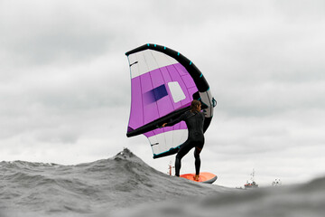 Surfer stands on a board in the waves, holding on to wings for wingfoiling © fesenko
