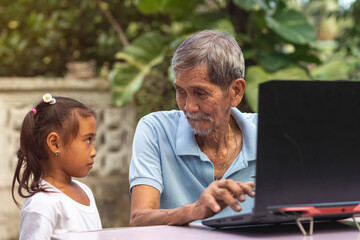 An old asian man in his 70s listens to his curious granddaughter while typing on a laptop while sitting outside at his garden.