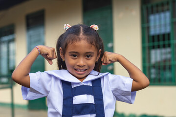 A confident preschooler asian lady flexes her arms. A little girl at a provincial primary school in Bohol. - obrazy, fototapety, plakaty