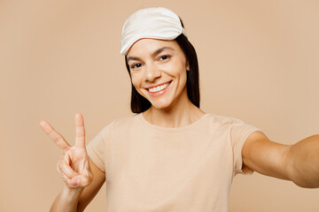 Close up young Latin woman wears pyjamas jam sleep eye mask rest relax at home do selfie shot on mobile cell phone show v-sign isolated on plain beige background studio. Good mood night nap concept.