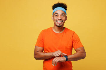 Young fitness trainer instructor sporty man sportsman wear orange t-shirt use smart watch to check calories spend time in home gym isolated on plain yellow background. Workout sport fit abs concept. - obrazy, fototapety, plakaty
