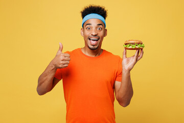 Young fitness trainer instructor sporty man sportsman wears orange t-shirt hold fastfood burger...