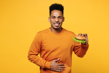 Young man wear sweatshirt casual clothes eat burger put hands abdomen suffer from stomach ache feel bad isolated on plain yellow background Proper nutrition healthy fast food unhealthy choice concept - obrazy, fototapety, plakaty
