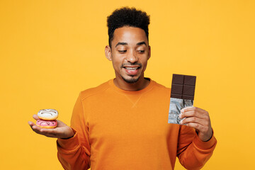 Young man wear orange sweatshirt casual clothes hold eat donut look at bar of chocolate isolated on plain yellow background studio portrait Proper nutrition healthy fast food unhealthy choice concept - obrazy, fototapety, plakaty