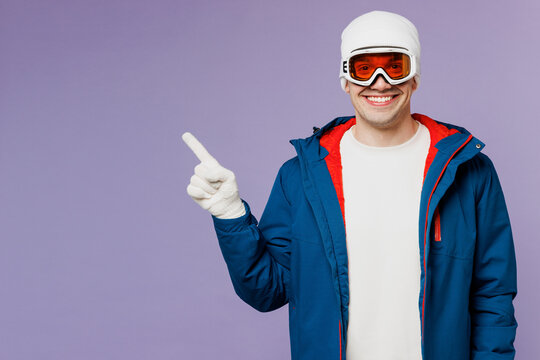 Skier smiling happy man wears warm blue windbreaker jacket ski goggles mask hat point index finger aside on area spend extreme weekend winter season in mountains isolated on plain purple background.