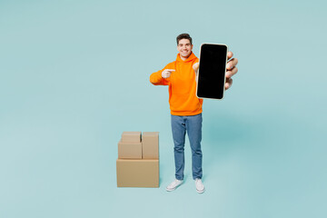 Full body young man wear orange hoody casual clothes stand near stack cardboard blank boxes point...
