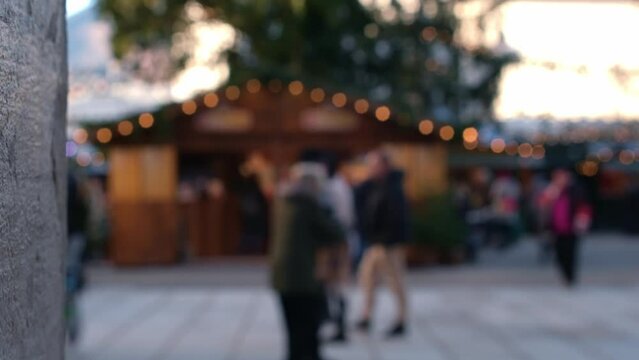 Blurred people walking through Christmas market. Trade tents with lights. Defocused people walk past shopping arcades. Illuminated fair. Background gradually becomes more blurred