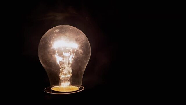 One Glowing Incandescent Light Bulb in Thick Smoke on Black Background. Close up. Dusty, switched-on Edison tungsten light bulb shines a yellow light in smoky dark room. Blurred motion. Texture. Fog.