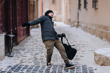 A young guy in a gray coat, hat and glove with a backpack walks along an old slippery snow-covered...