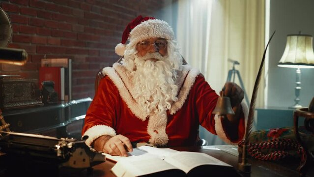 Portrait of Santa Claus looking at the camera and ringing a bell while sitting in his office at desk late at night on Christmas Eve