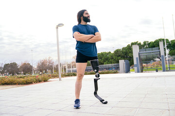 Smiling bearded sportsman with a running blade prosthesis stands in a park, embodying resilience...