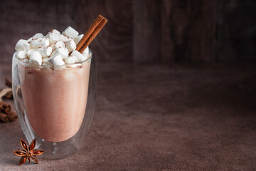 Sweet traditional hot chocolate or homemade cocoa drink decorated with marshmallow topping and cinnamon stick served in glass cup or mug on dark brown table with anise star spice. With copy space