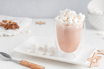 Seasonal sweet hot chocolate or cocoa drink decorated with marshmallow topping served in glass cup or mug on plate with ingredients on white wooden table with spoon and star shaped decoration 