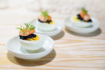 Party snacks from boiled eggs with caviar and shrimps on small white plates on a wooden table for a holiday buffet on Christmas, New Year or Easter, copy space, selected focus