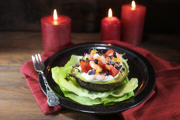 Halved avocado filled with shrimps, tomatoes and mayonnaise on a black plate, red candles and...